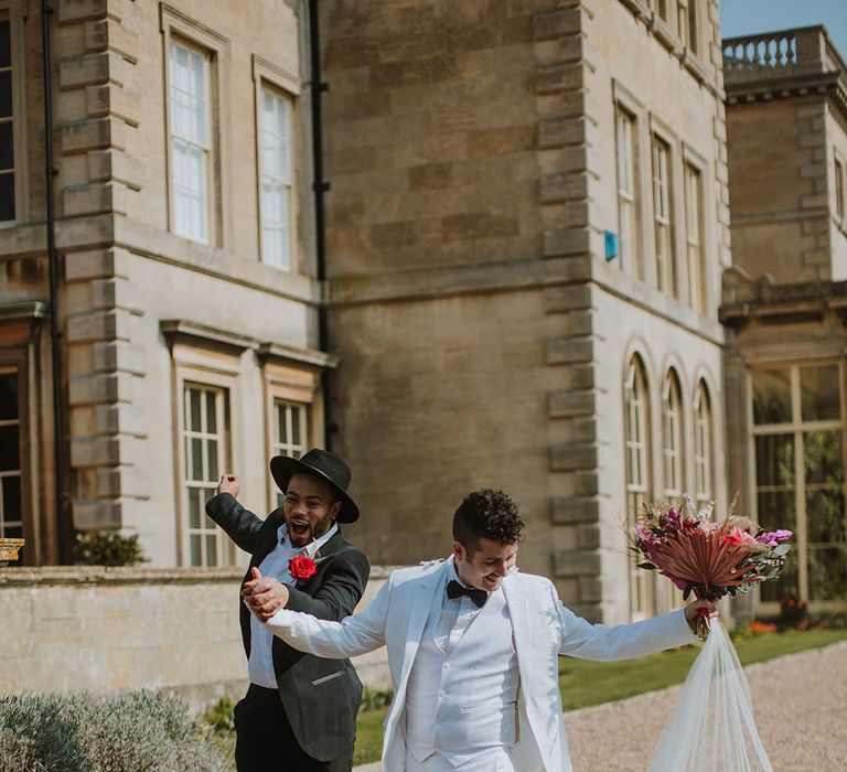 Groom in a two piece black suit, fedora hat and boots holding hands with his partner in a white three-piece groom suit, white boots and cape 