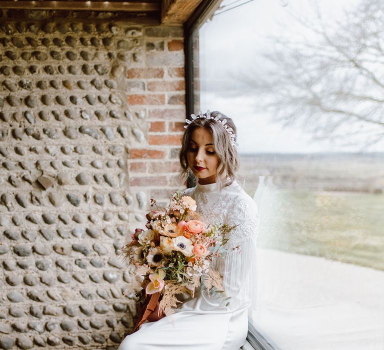 Bride in a satin wedding dress with fringe detail holding a blush pink bouquet sitting on the ledge at Montague Farm Hankham