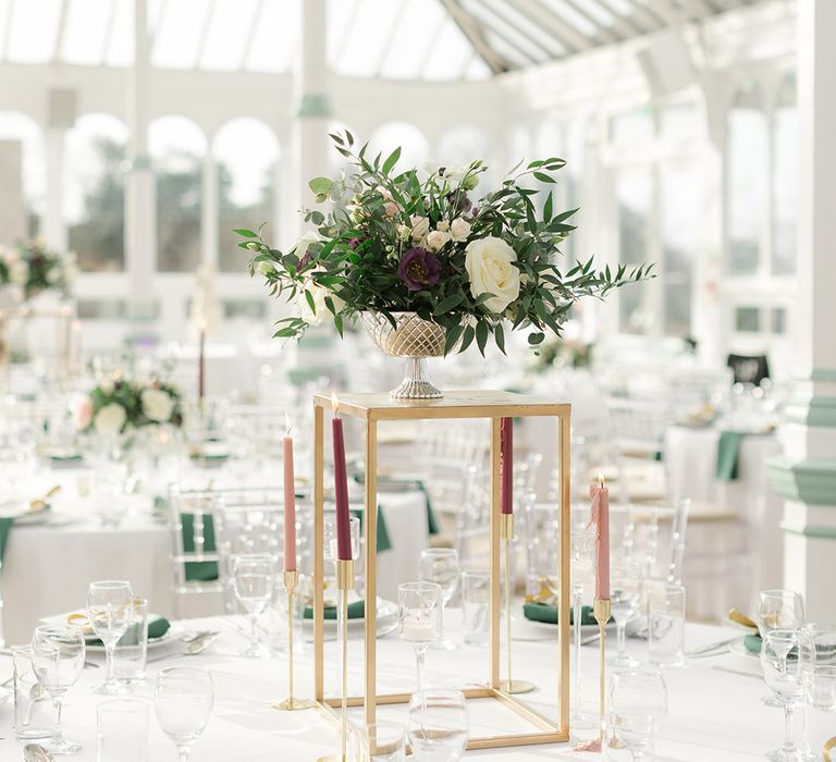 Floral bouquet sits in vase upon gold stand surrounded by candles in burgundy and pale pink 