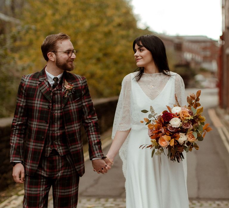 Bride who wears lace cape looks lovingly at her groom in bespoke Tartan suit as they walk to wedding reception