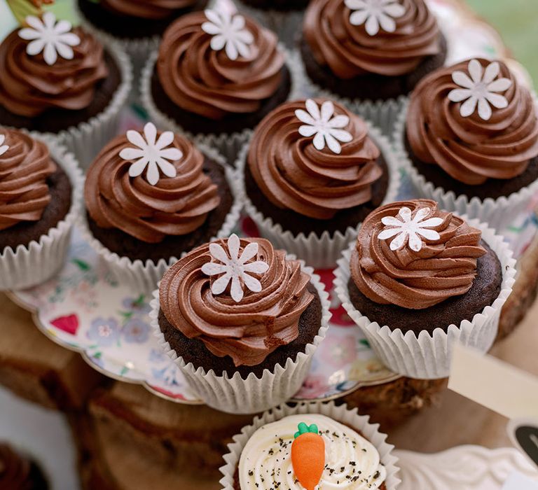 Chocolate frosted cupcakes complete with white flower next to carrot cake cupcakes complete with white frosting and small carrot 