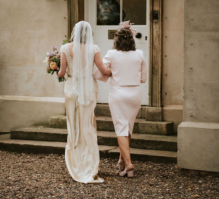 Bride in mid-length veil walks mother of the bride who wears pastel pink suit complete with fascinator 