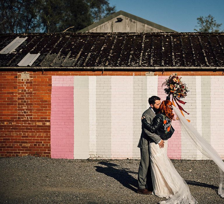 Bride and groom kiss in front of colourful wall with bride's veil flowing in the wind wearing personalised leather jacket and groom in a grey suit 