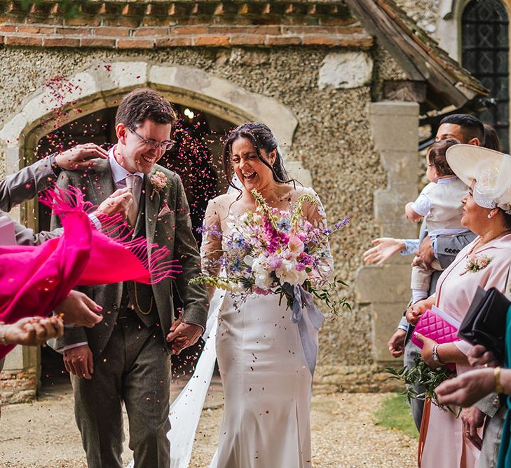 Bride holding tropical bouquet walks through colourful confetti outside church with her groom who wears grey three piece suit