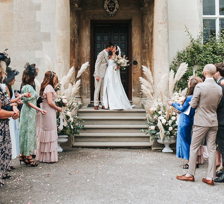 Bride and groom share a kiss at the entrance to Elmore Court decorated with pampas grass floral arrangements 