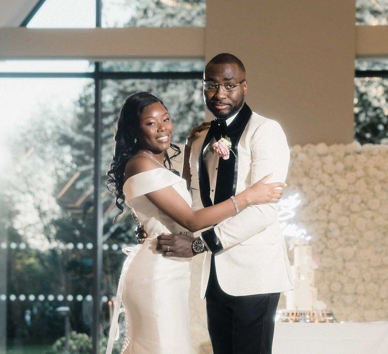 Bride wears fitted off-the-shoulder wedding dress finished with buttoned back and diamanté belt to the front as she stands beside her groom in black tie