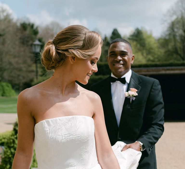 Bride with an intricate updo looks back at the groom as he lifts her wedding dress train as they walk 