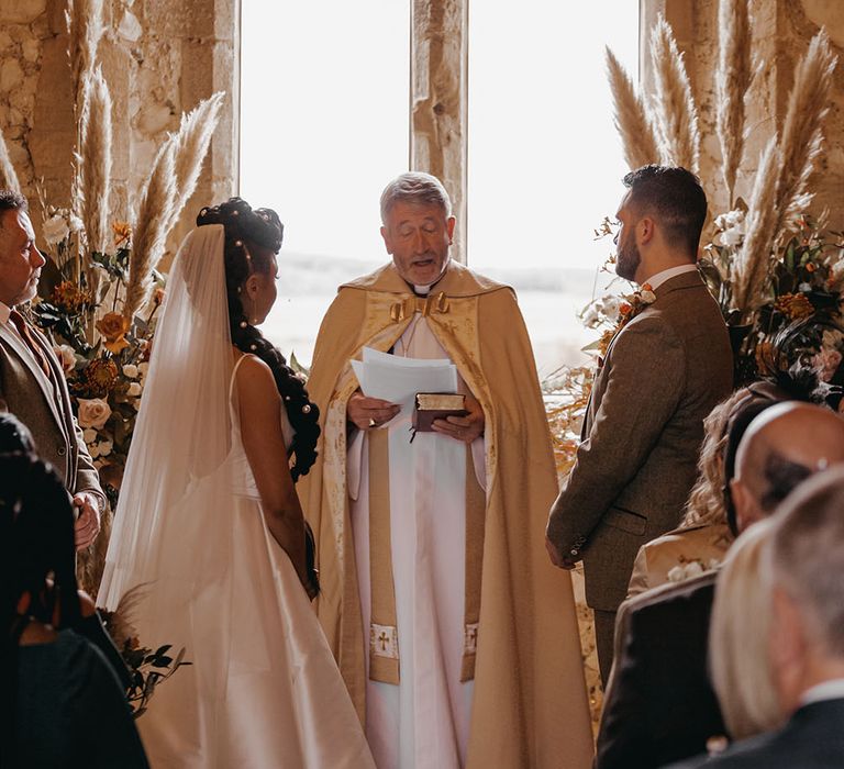 Bride & groom stand in front if Bishop during Christian wedding ceremony at Pentney Abbey surrounded by pampas grass arrangements with Autumnal colour scheme