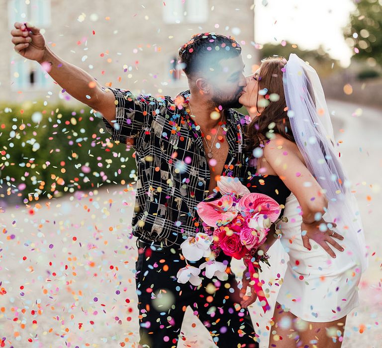 Groom pops colourful confetti around them as they kiss outdoors on their wedding day