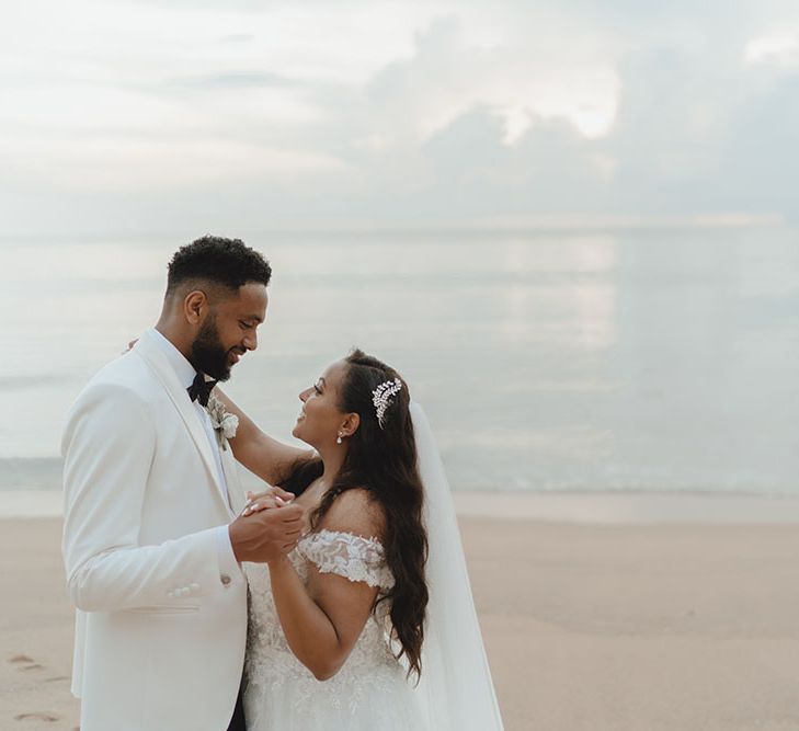 Bride wears floor-length veil and looks lovingly toward her groom on the seafront in Phuket 