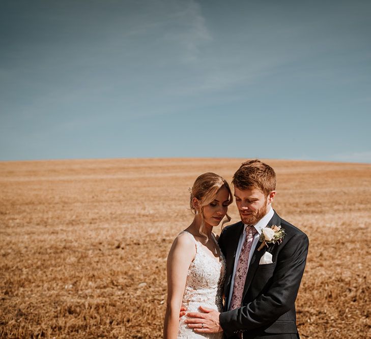 Groom wears floral buttonhole and stands beside his bride in golden fields 