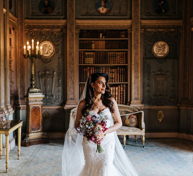 Indian bride wearing lace wedding dress with sweetheart neckline and wears her brown hair in loose waves whilst holding brightly coloured floral bouquet 