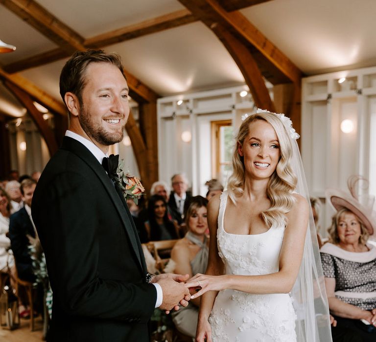 Groom in black tie puts the wedding ring on the bride's finger who has curled blonde hair with a floral veil and matching wedding dress