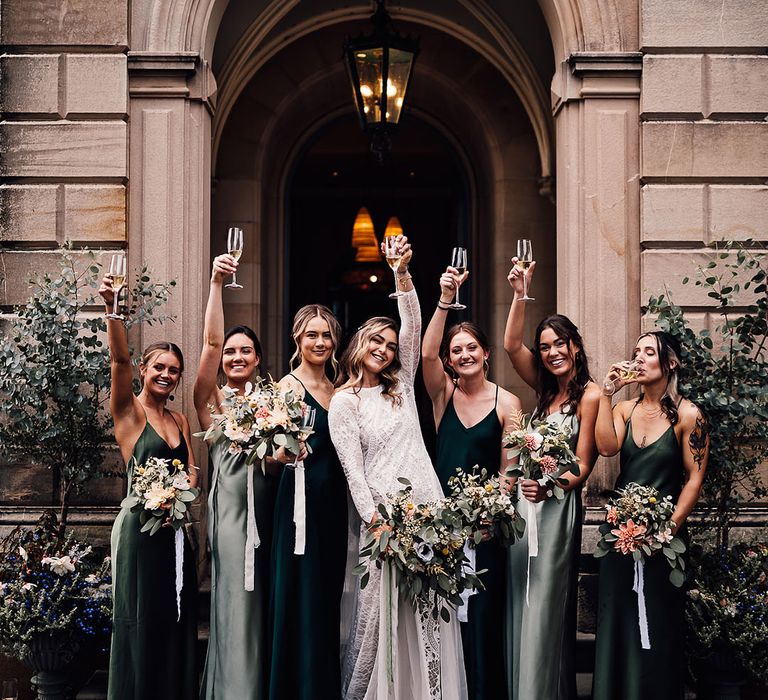 Bride in a long sleeve lace wedding dress stands outside of Callow Hall with her bridesmaids in different shades of green dresses drinking champagne