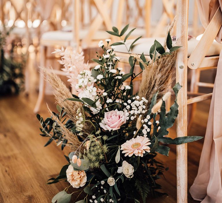 Neutral toned floral arrangement with green foliage beside chair lining the aisle 