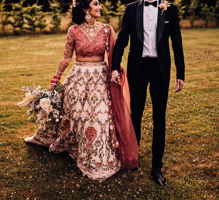 Indian bride wearing traditional lehenga walks with her groom outdoors in black tie 