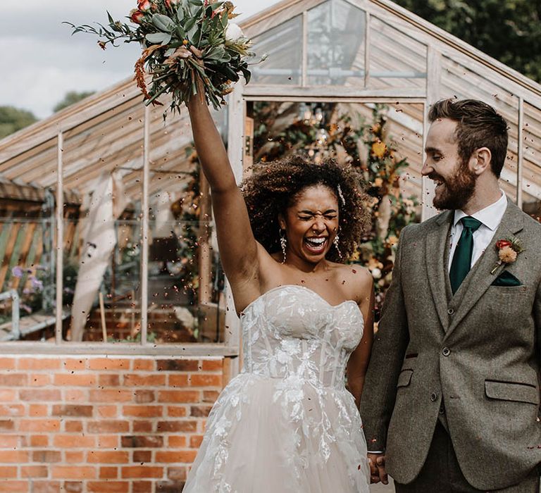 Bride in a strapless sparkly wedding dress and groom in a grey tweed suit have a confetti moment at their glasshouse reception