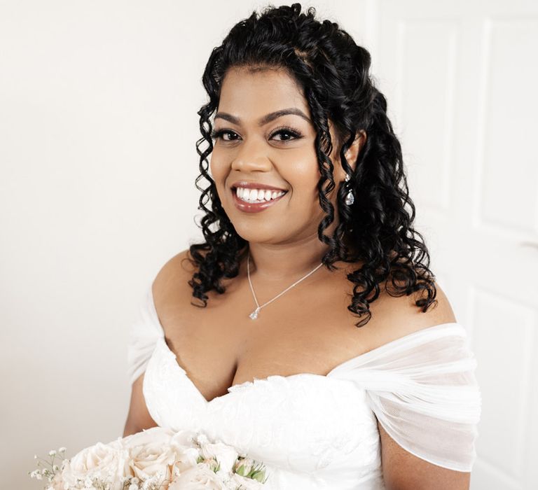 Bride wears her black hair in tight curls and wears off the shoulder sheer sleeved wedding dress with sweetheart neckline