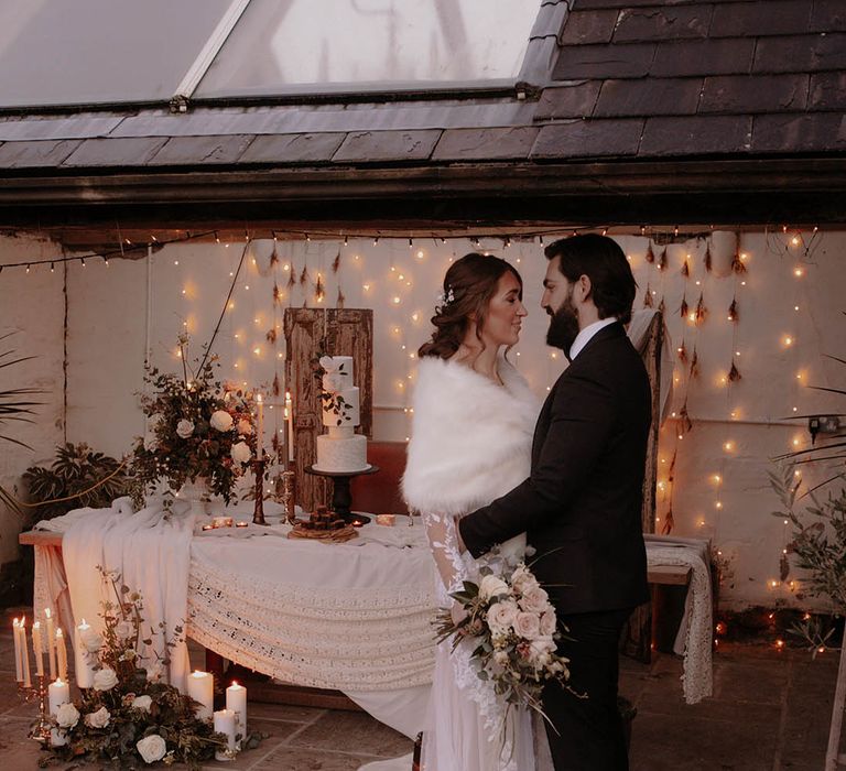 Rustic winter wedding tablescape with fairy lights, foliage and an iced winter wedding cake and layered table cloths with the groom in black tie and the bride in a lace wedding dress