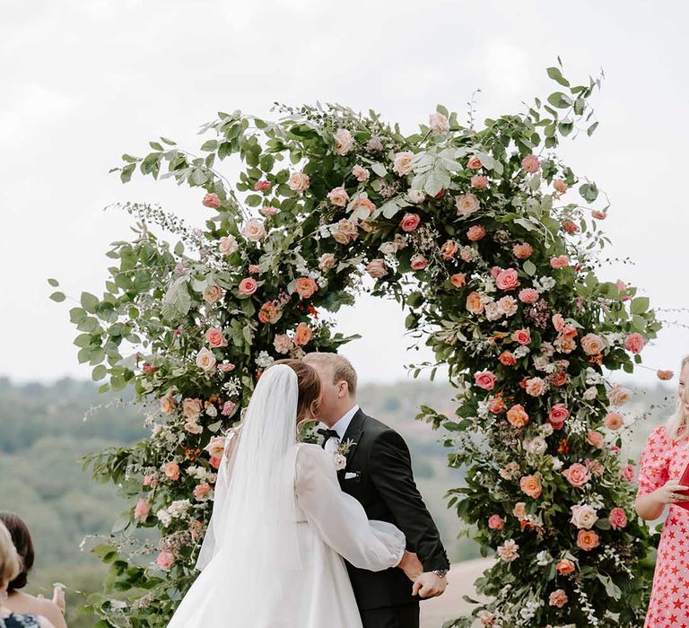 Bride wearing full sheer sleeve wedding dress kisses her groom in front of floral archway filled with orange and pink roses 