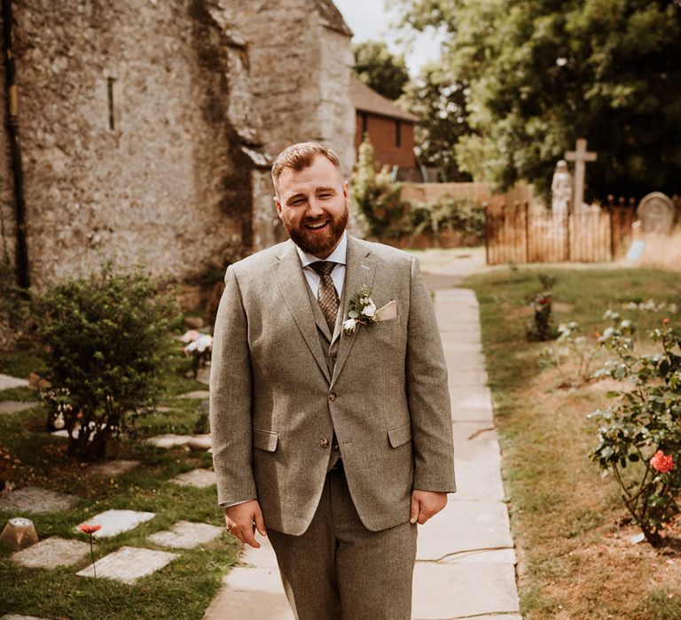 Bearded groom stands smiling in a three piece grey tweed suit 