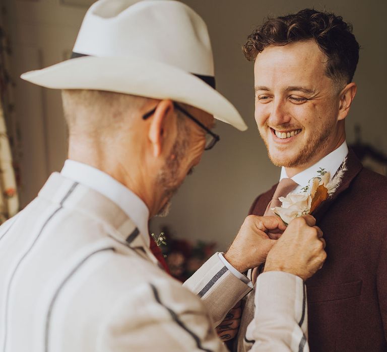 Groom has his buttonhole placed on maroon suit on the morning of his wedding day