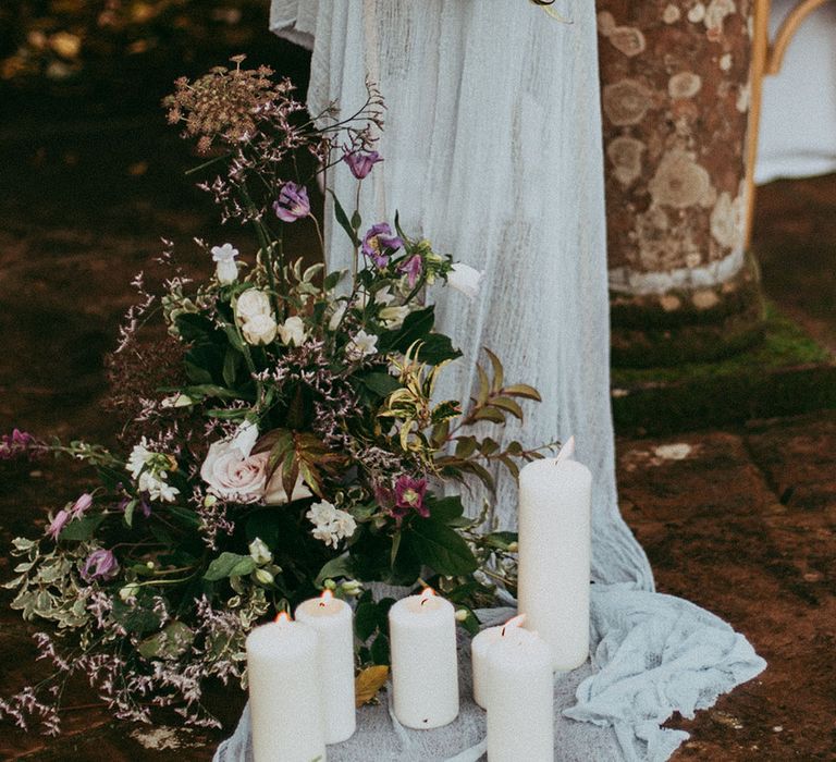 White pillar candles, sheer white drapes and mixed neutral floral decorations with white carnations, purple tulips, roses and foliage