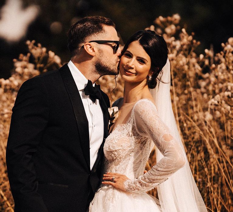 Groom in black tie with black sunglasses kisses the bride on the cheek for their cute couple portraits 