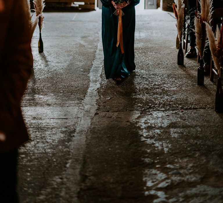 Bridesmaid walks down the aisle in green satin bridesmaid dress, carrying autumnal wedding flowers