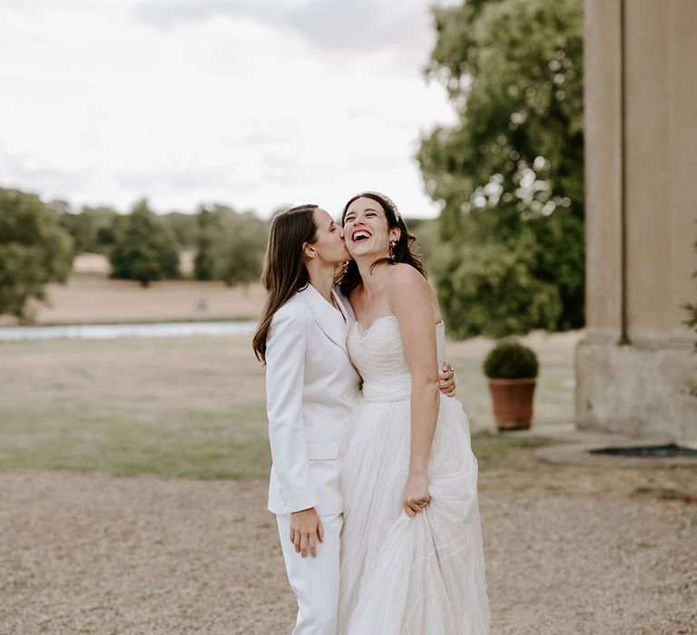 Bride in off the shoulder tulle wedding dress gets a kiss on the cheek from the bride in a white suit 