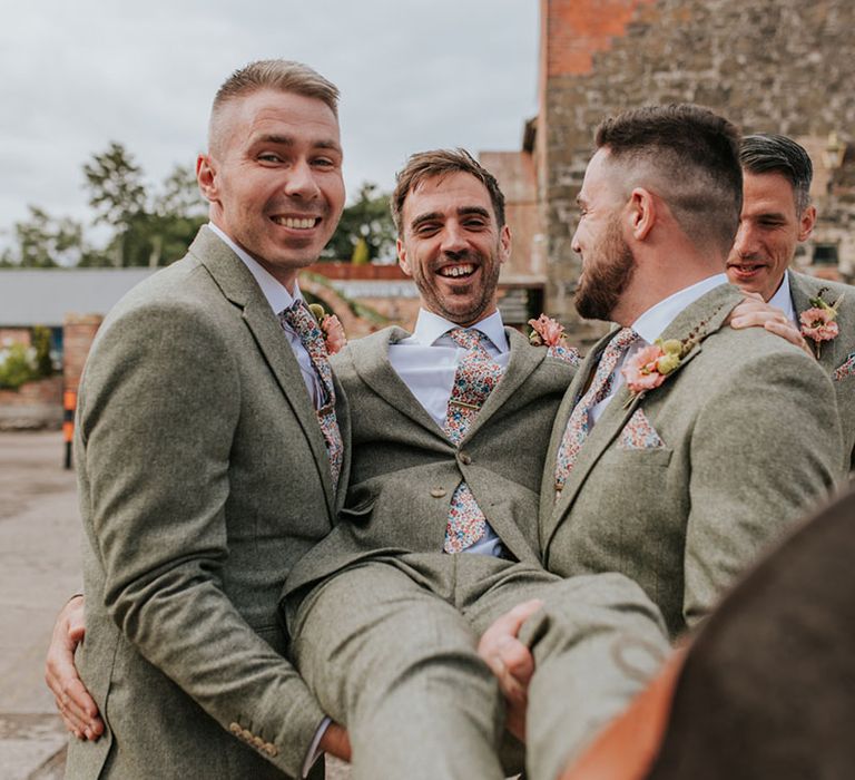 Groomsmen lift the groom as they all wear matching tweed suits with floral ties and pocket squares 