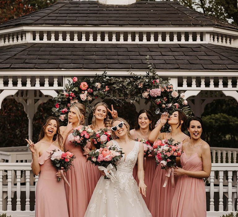 Bridesmaids in mismatched style pink dresses with the bride in front of a pink flower arch 