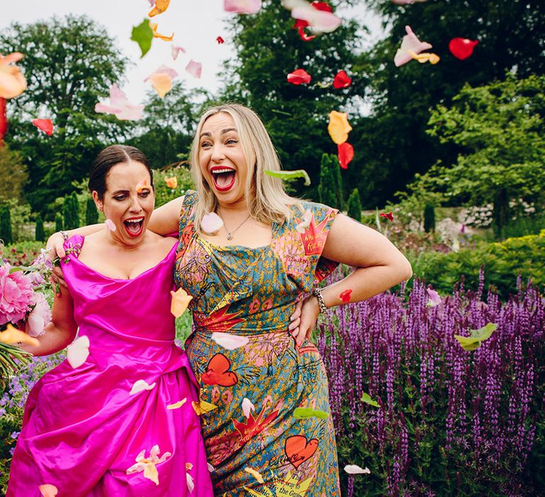 Bridesmaid in midi green colourful patterned Vivienne Westwood bridesmaid dress standing with bride in fuchsia off the shoulder Vivienne Westwood dress doing flower petal confetti throw