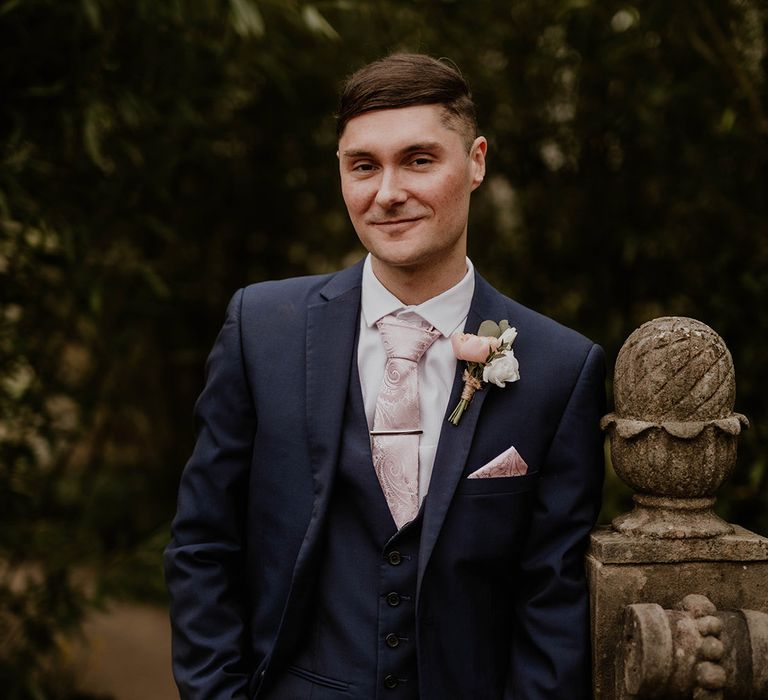 Groom wears three piece suit complete with pale pink tie, pocket square and floral buttonhole 