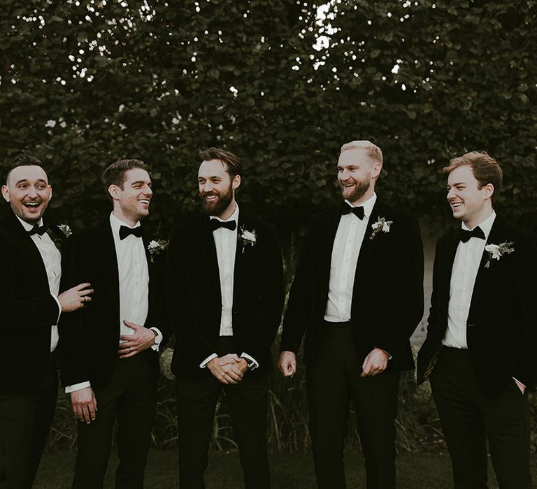 Groom and groomsmen stand laughing together in matching black tuxedos for the wedding day 