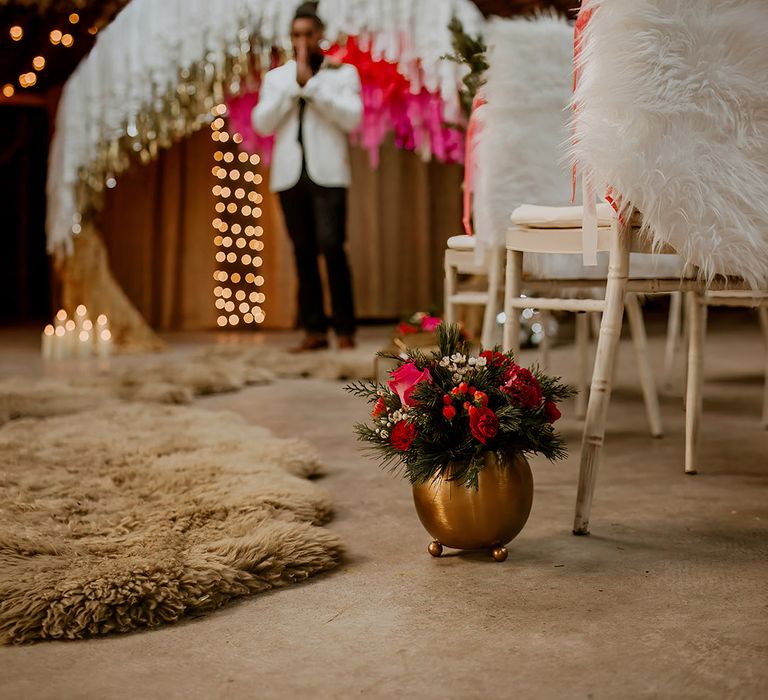 Groom doing first look at winter-styled boho wedding in white blazer standing at the end of the aisle with brass flower pots, faux fur throws down the aisle and on chairs, fairy lights and large semi-circle wedding arch with colourful streamers 