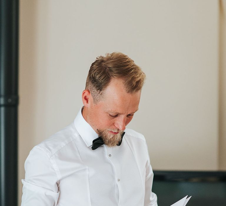 The groom in a white shirt with a black bow tie reading the wedding vows on the morning of the wedding 