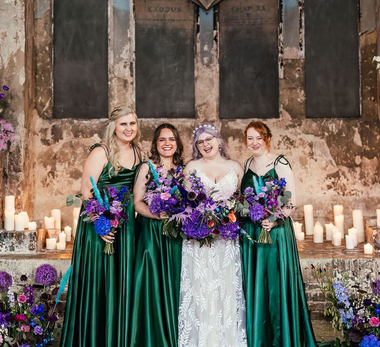 Bridesmaids in forest green satin bridesmaid dresses with tie straps and silver bejewelled celestial hair accessories standing with bride in dainty leaf 3d appliqué sheer bridal gown and cathedral length veil and celestial silver bridal headband, all holding bright purple bridal bouquets with purple hydrangeas, pink garden roses, purple orchids, lilac carnations, dried flowers and peacock blue feathers 