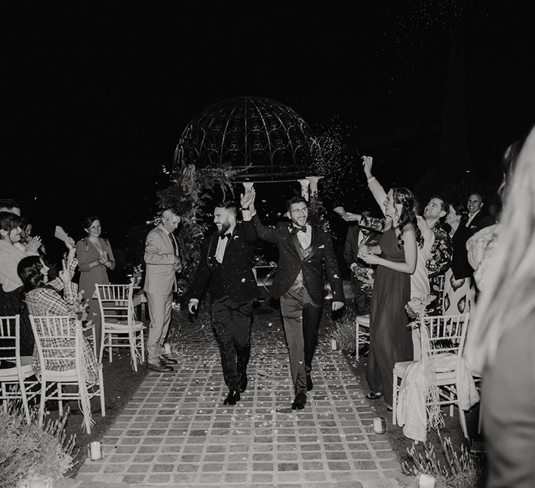 two grooms in tuxedos holding hands exiting their outdoor wedding ceremony 