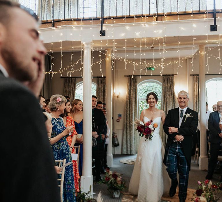 Father of the bride in tartan trousers walks his daughter down the aisle in a Stephanie Allin bridal gown with thin straps and polka dot tulle overlay 