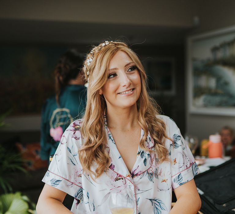 Bride with curled and braided hair with flower hair accessories sitting in floral satin pyjamas as she gets ready for the wedding 