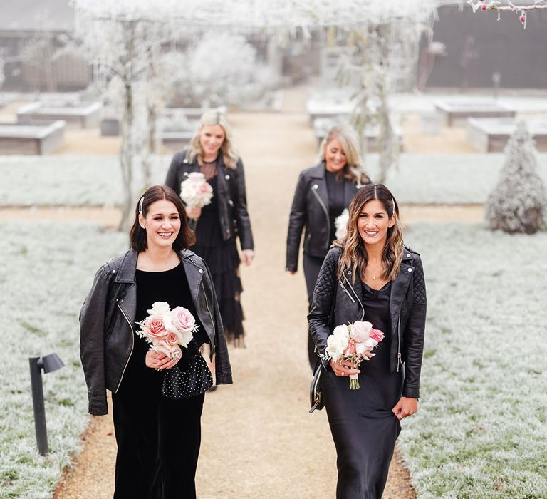 Bridesmaids in black bridesmaid dresses with matching leather jackets with small bouquets of pale pink roses 