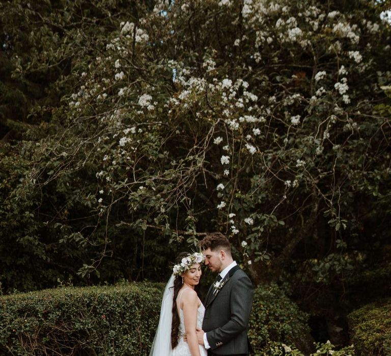 Bride wearing ethereal, celestial 3d appliqué sheer overlay wedding dress and bridal flower crown holding large white rose, peony, dried flower and foliage bridal bouquet embracing groom in dark grey blazer and waistcoat, dark tartan tie with white flower boutonniere, dark grey and blue tartan kilt and wedding sporran 