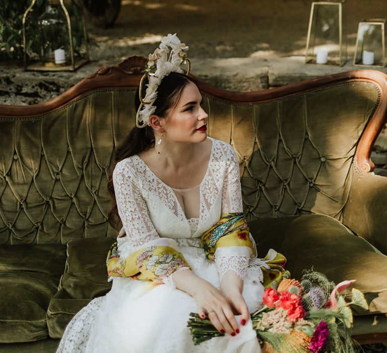 Bride in lace wedding dress and bridal crown with bridal bouquet consisting of red pincushion flowers, coral carnations, magenta peonies and anthuriums, sitting on a vintage olive green upholstered sofa