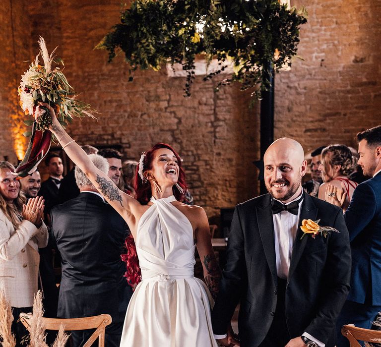 Bride with long red hair in halter neck wedding dress walks back down the aisle with the groom in black tie as a married couple 