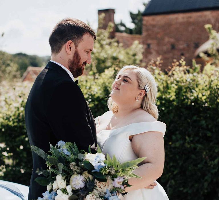 Groom wearing classic black grooms three piece tuxedo with white rose boutonniere and black bowtie embracing bride in satin off the shoulder wedding dress holding white and blue eucalyptus, foliage, garden rose, gardenia, light blue delphinium and dried flower bridal bouquet tied with dusky blue ribbon at Brickhouse Vineyard Devon wedding