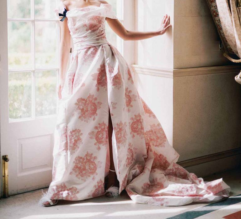 Bride in off the shoulder white and pink floral patterned bridal gown with black bow and front slit standing by window at Settrington Orangery