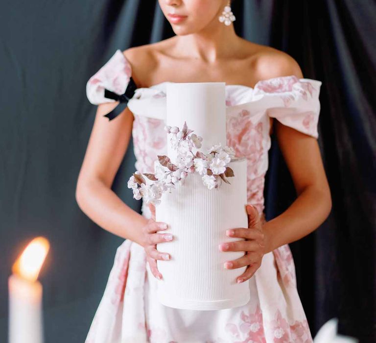 Bride in off the shoulder white and pink floral patterned bridal gown with black bow and front slit holding two tiered iced buttercream wedding cake with spring flower decorations 