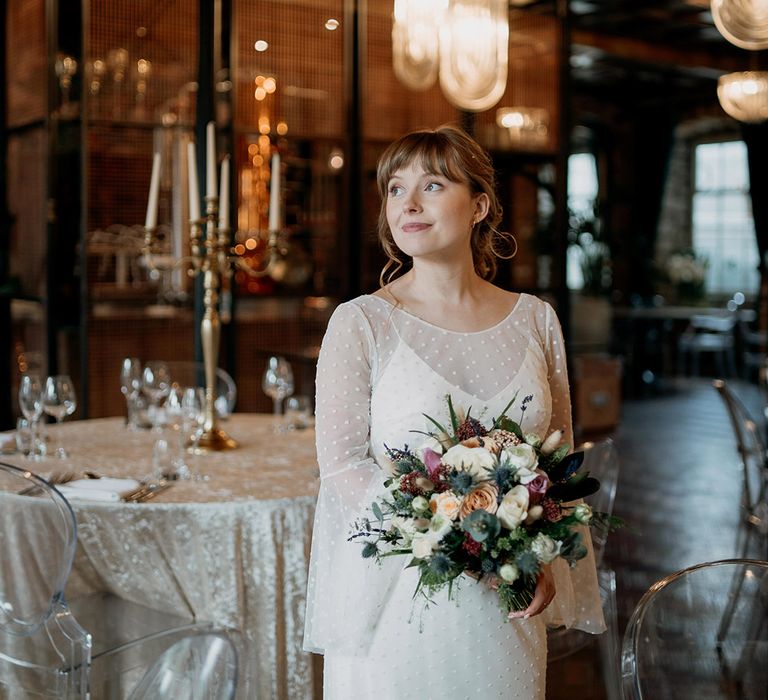 The bride wears a long sleeve dotted wedding dress with veil holding purple, red, and white wedding bouquet 