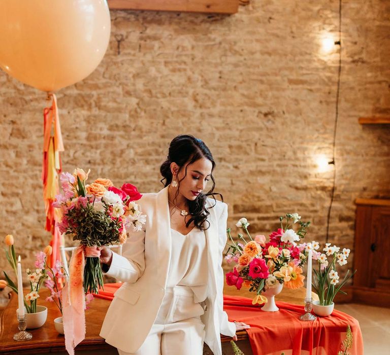 Bride in white bridal suit with closed toe bright green wedding heels standing by wooden table with orange table runner and mixed seasonal flower arrangements at Merriscourt wedding venue